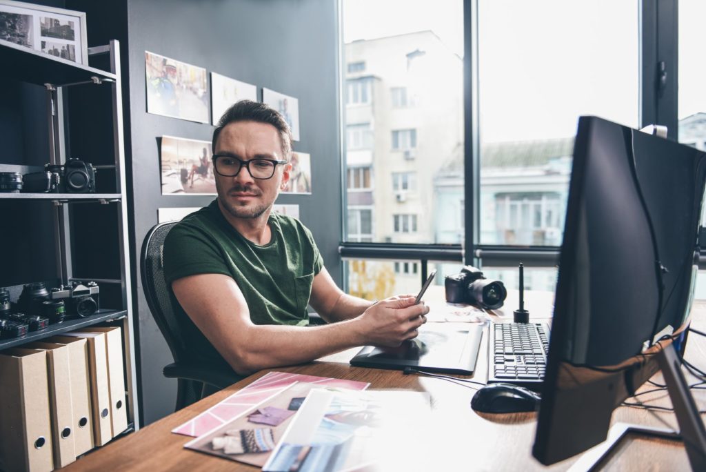 Portrait-of-smiling-unshaven-man-keeping-mobile-while-locating-at-desk-with-digital-device-cameras-situating-in-office | OnTrack